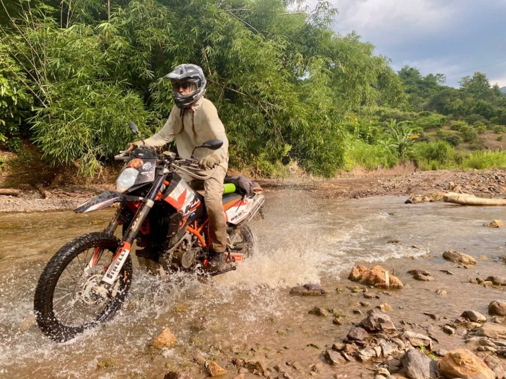 Motorcyclist riding an off-road bike through a shallow river in a lush, tropical landscape